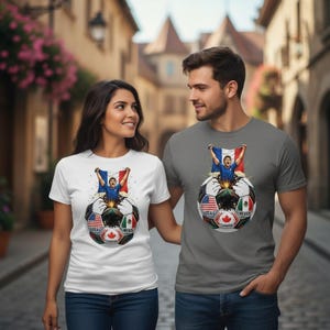 Op de afbeelding: Een man en een vrouw dragen t-shirts met een voetbalontwerp. De bal toont vlaggen van de VS, Mexico en Canada. Een persoon houdt een vlag boven de bal. Het shirt van de vrouw is wit en dat van de man grijs.