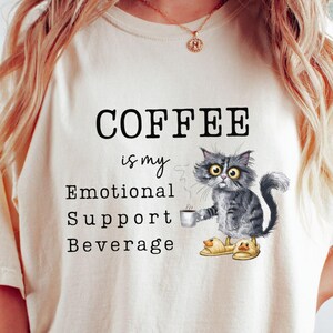 May include: Ivory t-shirt with the text "COFFEE is my Emotional Support Beverage." A watercolour illustration of a grey cat holding a coffee cup is featured below the text. The shirt is a casual, comfortable style.