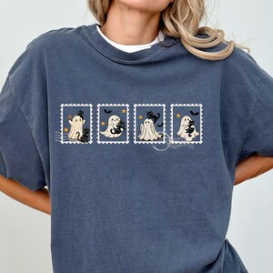 May include: Denim blue t-shirt with a white crew neck. The shirt features four postage stamp designs with cartoon ghosts, bats, and stars. The word "DENIM" is printed in white at the bottom of the shirt.