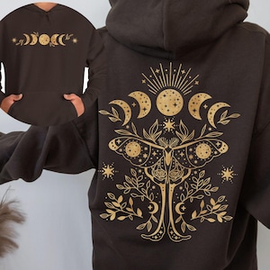 May include: A dark brown hoodie with a gold celestial moth and moon phase design on the back. The front features a smaller moon phase design. The hoodie has a hood and a front pocket.
