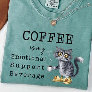 May include: Light green t-shirt featuring the text "COFFEE is my Emotional Support Beverage" and a cartoon cat with a coffee cup. The cat wears duck slippers. A gold necklace is draped across the shirt.