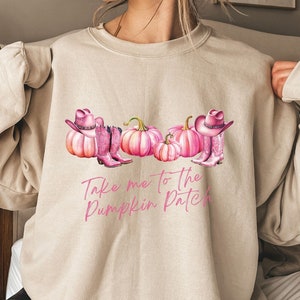 May include: Beige sweatshirt featuring a graphic of pink pumpkins, cowboy boots, and hats. The text "Take me to the Pumpkin Patch" is written in pink script. The sweatshirt is a neutral colour.