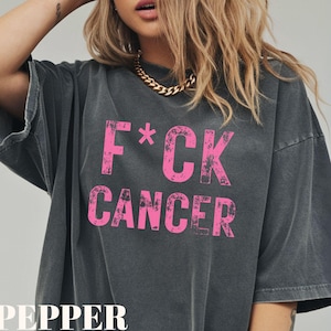 May include: Dark gray oversized t-shirt with the text "F*CK CANCER" in bright pink. The word "PEPPER" is printed in white at the bottom of the shirt. The shirt has short sleeves and a relaxed fit.