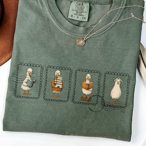 May include: Moss green t-shirt with a crew neck and short sleeves. The shirt features a design of four ducks in postage stamp frames, each with a different book-related activity. A brown hat and necklace are also visible.
