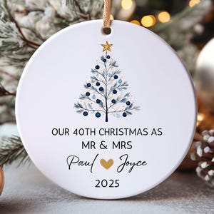 May include: A white ceramic Christmas ornament with a gold string. The ornament features a blue and white Christmas tree design and the text "OUR 40TH CHRISTMAS AS MR & MRS Paul ❤ Joyce 2025."