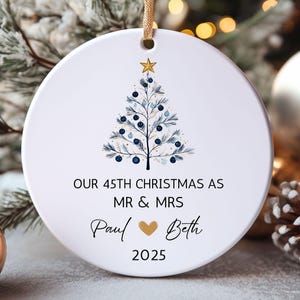 May include: A white ceramic Christmas ornament with a gold string. The ornament features a blue and gold Christmas tree design and the text "OUR 45TH CHRISTMAS AS MR & MRS Paul ❤ Beth 2025". A gold ball and pinecones are in the background.
