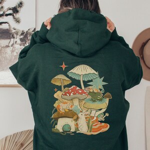 May include: A dark green hooded sweatshirt with a colorful mushroom graphic on the back. The graphic features a variety of mushrooms, including a red and white mushroom with white spots, a blue and green mushroom, and a yellow mushroom. There is also a small white rabbit and a frog in the graphic.