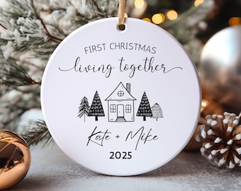 May include: A white ceramic Christmas ornament with the text "First Christmas living together" and a house illustration. The names "Kate + Mike" and the year "2025" are also printed on the ornament. The ornament hangs from a gold string.