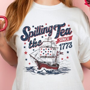 May include: White t-shirt with a vintage-style graphic of a ship sailing on the ocean. The graphic includes the text "Spilling the Tea Since 1773" with stars and red, white, and blue accents.