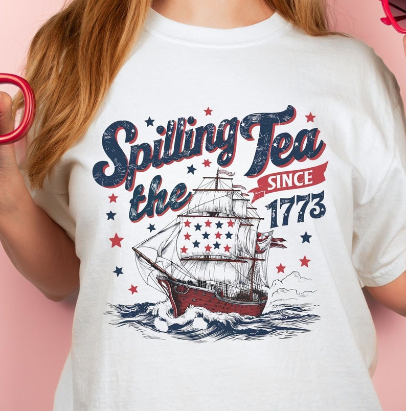 May include: White t-shirt with a vintage-style graphic of a ship sailing on the ocean. The graphic includes the text "Spilling the Tea Since 1773" with stars and red, white, and blue accents.