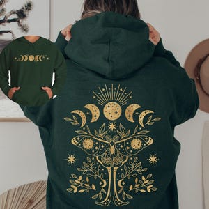 May include: Dark green hoodie with a gold celestial design on the back, featuring a sun, moons, and a moth. The front of the hoodie has a smaller gold design of moon phases. A hat hangs on the wall in the background.
