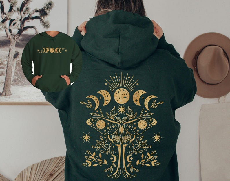 May include: Dark green hoodie with a gold celestial design on the back, featuring a sun, moons, and a moth. The front of the hoodie has a smaller gold design of moon phases. A hat hangs on the wall in the background.