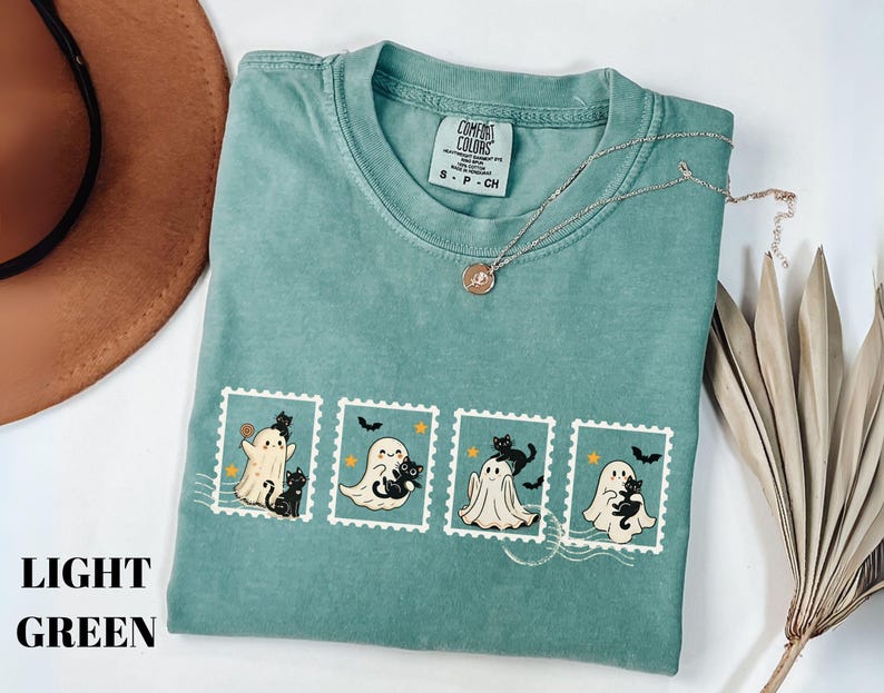 May include: Light green t-shirt featuring a Halloween-themed design. The design includes four postage stamp illustrations with ghosts, black cats, and bats. A gold necklace is draped across the shirt. The shirt is folded and placed on a white surface.
