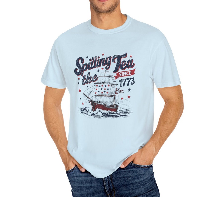 May include: A light blue t-shirt with a vintage-style graphic of a ship sailing on the ocean. The graphic includes the text "Spilling the Tea Since 1773" and stars.