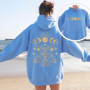 May include: A light blue hoodie with a gold celestial design on the back and front. The design includes phases of the moon, a sun, stars, and a tree. The hoodie is made of a soft material and has a hood and a front pocket.