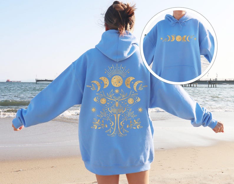 May include: A light blue hoodie with a gold celestial design on the back and front. The design includes phases of the moon, a sun, stars, and a tree. The hoodie is made of a soft material and has a hood and a front pocket.
