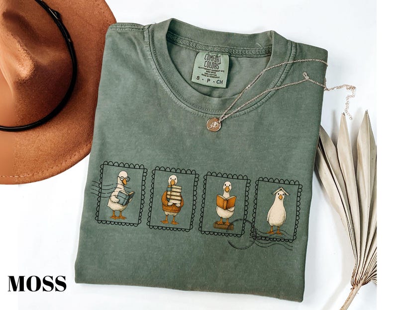 May include: Moss green t-shirt with a crew neck and short sleeves. The shirt features a design of four ducks in postage stamp frames, each with a different book-related activity. A brown hat and necklace are also visible.