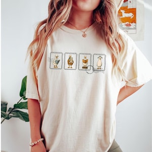 May include: Cream-colored t-shirt featuring four postage stamp designs with cartoon characters. Each stamp depicts a different character interacting with books, with a postmark detail. The shirt is a casual, comfortable style.