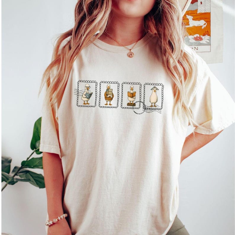 May include: Cream-colored t-shirt featuring four postage stamp designs with cartoon characters. Each stamp depicts a different character interacting with books, with a postmark detail. The shirt is a casual, comfortable style.