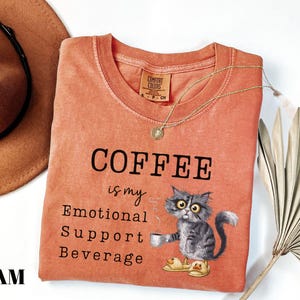May include: Orange Comfort Colors t-shirt with the text "COFFEE is my Emotional Support Beverage." A cartoon cat wearing slippers and holding a coffee cup is printed on the shirt. A gold necklace is draped across the shirt.
