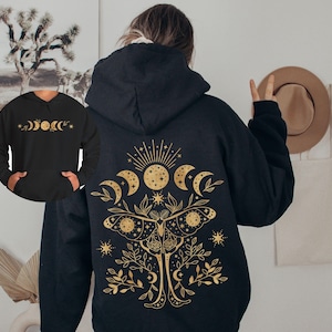 May include: Black hoodie with a gold celestial design on the back, featuring moons, stars, and a moth. The front of the hoodie has a smaller gold design of moon phases. The hoodie has a hood and a front pocket.
