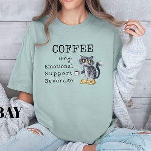 May include: A pale green t-shirt featuring the phrase "COFFEE is my Emotional Support Beverage." A cartoon cat wearing boots and holding a coffee cup is printed on the shirt. The word "BAY" is printed in the lower left corner.