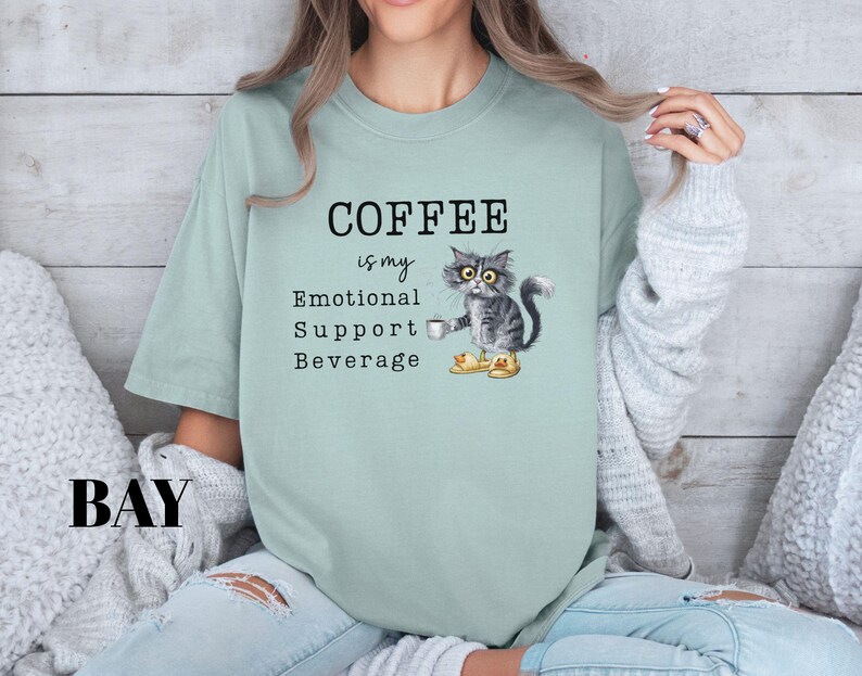 May include: A pale green t-shirt featuring the phrase "COFFEE is my Emotional Support Beverage." A cartoon cat wearing boots and holding a coffee cup is printed on the shirt. The word "BAY" is printed in the lower left corner.
