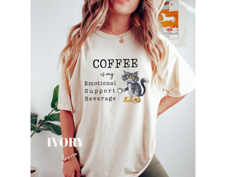 May include: Ivory t-shirt with the text "COFFEE is my Emotional Support Beverage." A watercolour illustration of a grey cat holding a coffee cup is featured below the text. The shirt is a casual, comfortable style.