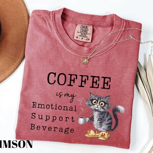 May include: Crimson-coloured t-shirt with the text "COFFEE is my Emotional Support Beverage." A cartoon cat wearing duck slippers holds a coffee cup. A gold necklace and a brown hat are also in the image.