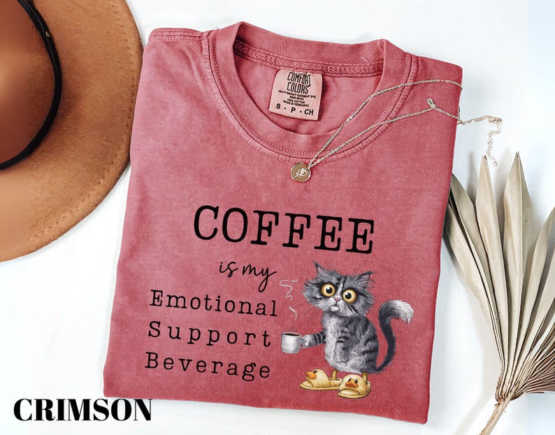 May include: Crimson-coloured t-shirt with the text "COFFEE is my Emotional Support Beverage." A cartoon cat wearing duck slippers holds a coffee cup. A gold necklace and a brown hat are also in the image.