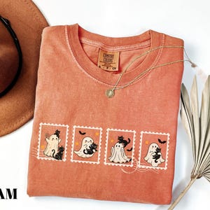 May include: A coral-colored t-shirt featuring a graphic of four postage stamps with Halloween-themed illustrations. Each stamp depicts a ghost and a black cat. A gold necklace and a brown hat are also visible.