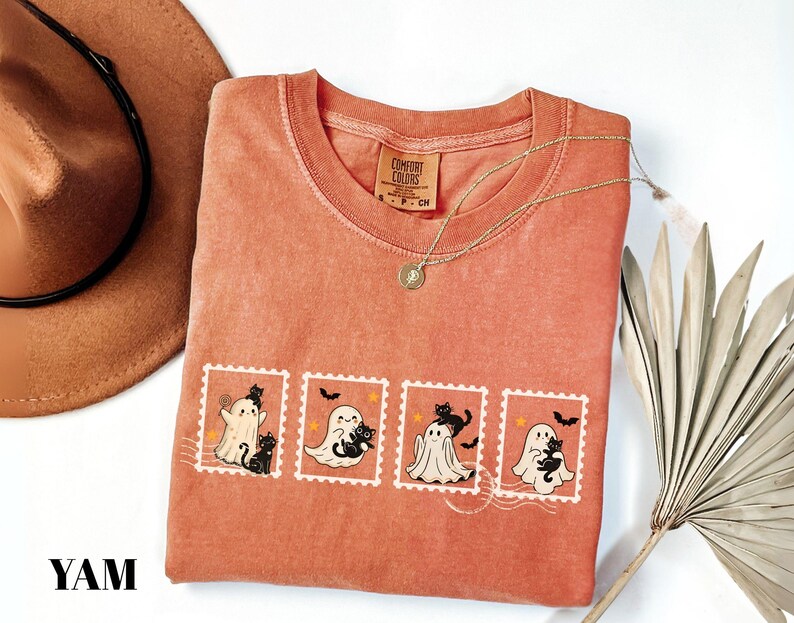 May include: A coral-colored t-shirt featuring a graphic of four postage stamps with Halloween-themed illustrations. Each stamp depicts a ghost and a black cat. A gold necklace and a brown hat are also visible.