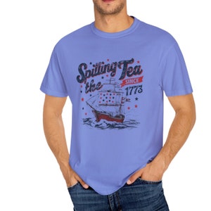 May include: A light blue t-shirt with a vintage-style graphic of a ship sailing on the water. The graphic includes the text "Spilling Tea the Since 1773".