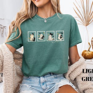 May include: Light green t-shirt featuring four postage stamp designs with cartoon ghosts and black cats. The shirt has short sleeves and a crew neck. The words "LIGHT GREEN" are printed in a stacked format on the bottom right.