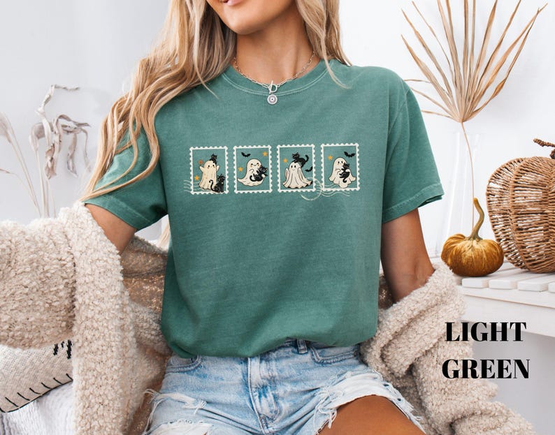May include: Light green t-shirt featuring four postage stamp designs with cartoon ghosts and black cats. The shirt has short sleeves and a crew neck. The words "LIGHT GREEN" are printed in a stacked format on the bottom right.