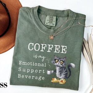 May include: Moss green t-shirt featuring the text "COFFEE is my Emotional Support Beverage" and a cartoon cat illustration. The cat is holding a coffee cup and wearing slippers. A brown hat and necklace are also visible.