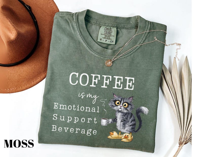 May include: Moss green t-shirt featuring the text "COFFEE is my Emotional Support Beverage" and a cartoon cat illustration. The cat is holding a coffee cup and wearing slippers. A brown hat and necklace are also visible.