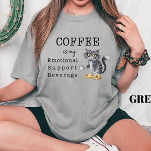 May include: Grey t-shirt featuring the text "COFFEE is my Emotional Support Beverage" and a graphic of a cat holding a coffee cup. The cat is wearing yellow slippers. The word "GREY" is printed in black.