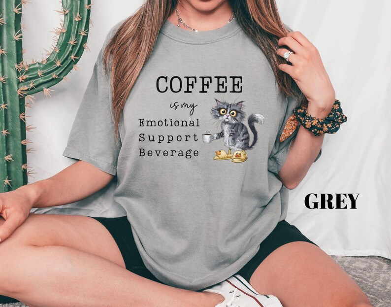 May include: Grey t-shirt featuring the text "COFFEE is my Emotional Support Beverage" and a graphic of a cat holding a coffee cup. The cat is wearing yellow slippers. The word "GREY" is printed in black.