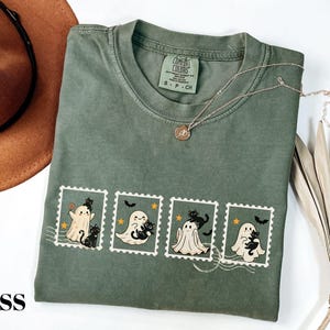 May include: Moss green t-shirt featuring a stamp design with cartoon ghosts and black cats. The shirt has a crew neck and short sleeves. A gold necklace and a brown hat are also in the image.