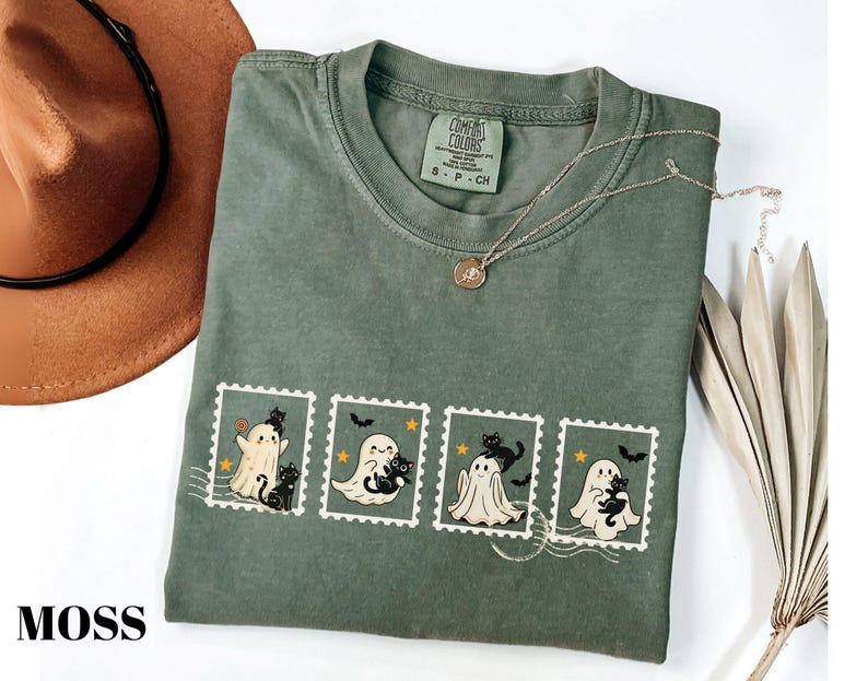 May include: Moss green t-shirt featuring a stamp design with cartoon ghosts and black cats. The shirt has a crew neck and short sleeves. A gold necklace and a brown hat are also in the image.