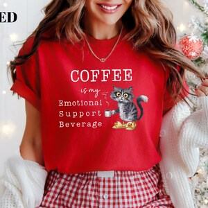 May include: Red t-shirt featuring the text "COFFEE is my Emotional Support Beverage" and a cartoon cat illustration. The cat is grey and white with yellow eyes, holding a coffee cup. The shirt has short sleeves.