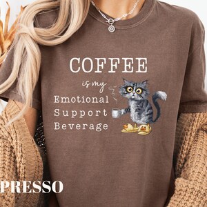 May include: A brown t-shirt with the slogan "COFFEE is my Emotional Support Beverage." A cartoon cat wearing duck slippers is depicted holding a coffee cup. The shirt is a casual style.