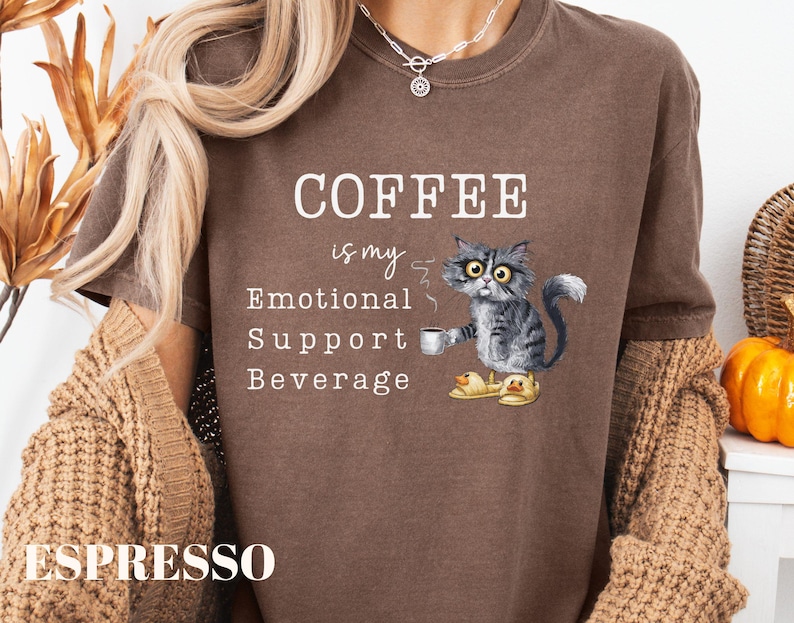 May include: A brown t-shirt with the slogan "COFFEE is my Emotional Support Beverage." A cartoon cat wearing duck slippers is depicted holding a coffee cup. The shirt is a casual style.