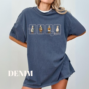 May include: Denim blue t-shirt with a white crew neck collar. The shirt features a design of four ducks in postage stamp frames. The word "DENIM" is printed in white at the bottom of the shirt.