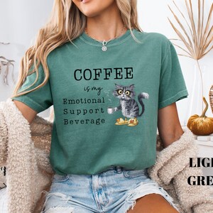 May include: Light green t-shirt featuring the text "COFFEE is my Emotional Support Beverage." A grey cat wearing glasses and slippers is holding a coffee cup. The words "LIGHT GREEN" are at the bottom of the shirt.