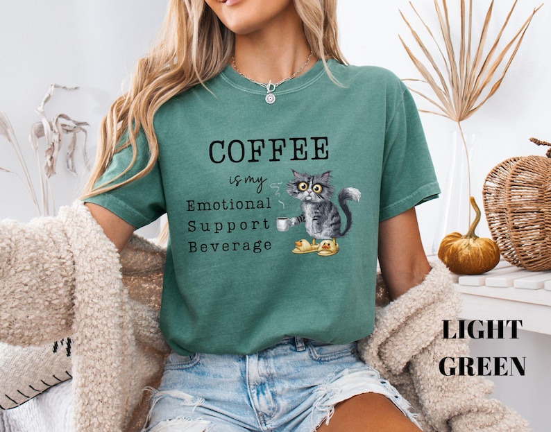 May include: Light green t-shirt featuring the text "COFFEE is my Emotional Support Beverage." A grey cat wearing glasses and slippers is holding a coffee cup. The words "LIGHT GREEN" are at the bottom of the shirt.