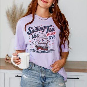 May include: A lavender t-shirt with a vintage-style graphic of a ship with the text "Spilling the Tea Since 1773".
