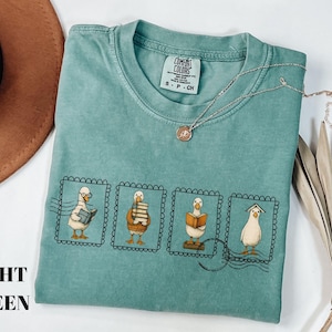 May include: Light green t-shirt with a graphic of four geese reading books, each in a postage stamp frame. The shirt has a crew neck and short sleeves. A gold necklace with a pendant is on the shirt. The words "LIGHT GREEN" are printed on the left side.