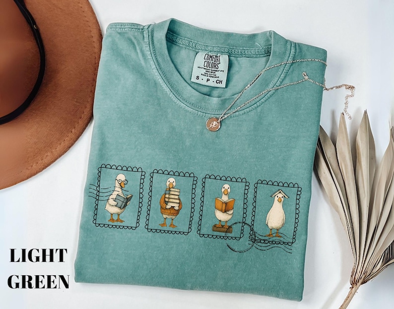 May include: Light green t-shirt with a graphic of four geese reading books, each in a postage stamp frame. The shirt has a crew neck and short sleeves. A gold necklace with a pendant is on the shirt. The words "LIGHT GREEN" are printed on the left side.